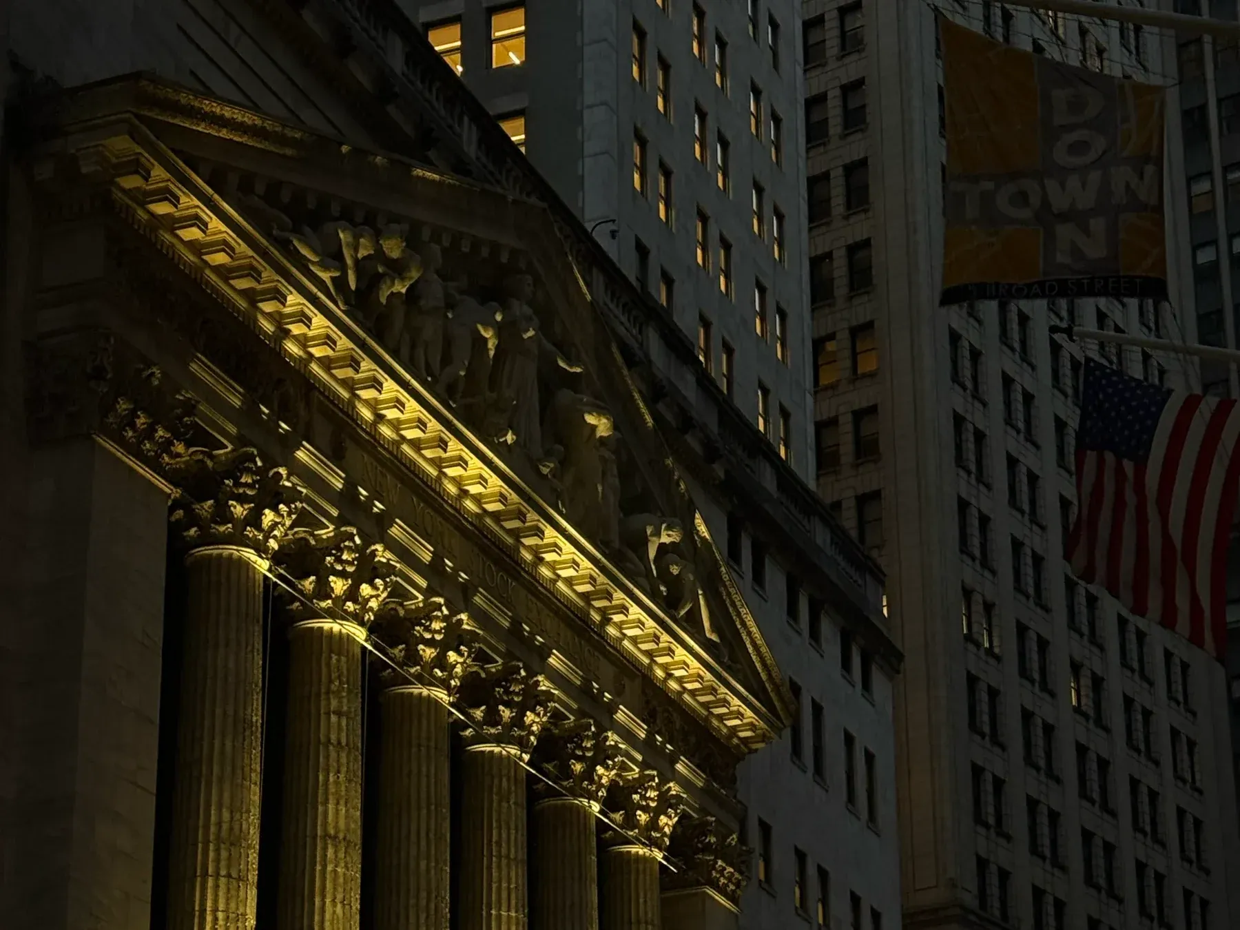 The New York Stock Exchange lit at night from Broad Street, with a Downtown banner and a flag on the building opposite